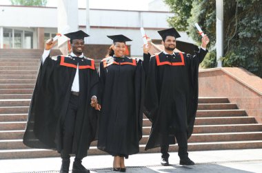 Mezuniyet törenine katılan çeşitli uluslararası öğrenciler mezuniyet şapkası ve cüppeli mutlu çok ırklı öğrenciler üniversite kampüsünde sıraya dizilmiş.