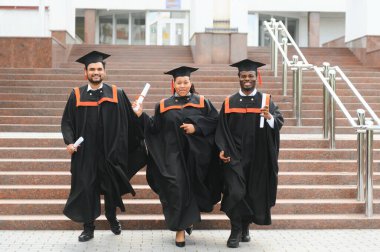 Mezuniyet törenine katılan çeşitli uluslararası öğrenciler mezuniyet şapkası ve cüppeli mutlu çok ırklı öğrenciler üniversite kampüsünde sıraya dizilmiş.