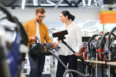 Çömelmiş çiftler ve satın almak istedikleri elektrik süpürgesine bakıyorlar. Teknoloji mağazası içi.