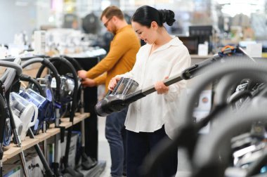 Çömelmiş çiftler ve satın almak istedikleri elektrik süpürgesine bakıyorlar. Teknoloji mağazası içi.