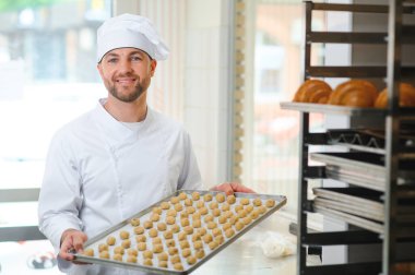 İyi kalitede. Steril giysi parlak gıda fabrikasında ayakta olgun neşeli erkek çalışanın resmini. Tepsi ile taze kurabiye dolu tutarak ve kalitesi iyidir eliyle işaret.
