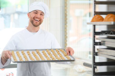 İyi kalitede. Steril giysi parlak gıda fabrikasında ayakta olgun neşeli erkek çalışanın resmini. Tepsi ile taze kurabiye dolu tutarak ve kalitesi iyidir eliyle işaret.