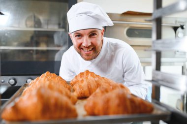 Genç, yakışıklı bir fırıncı elinde Fransız kruvasanı ile bir tepsi tutuyor ve gülümsüyor..