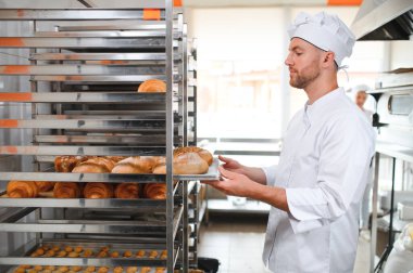 Male baker holding board with assortment of fresh bread in shop.
