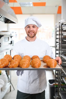 Genç, yakışıklı bir fırıncı elinde Fransız kruvasanı ile bir tepsi tutuyor ve gülümsüyor..