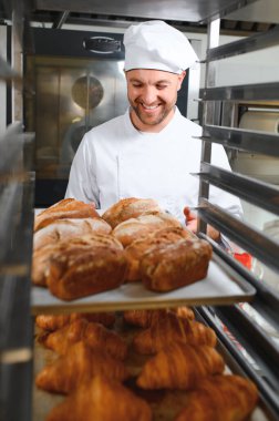 Male baker holding board with assortment of fresh bread in shop.