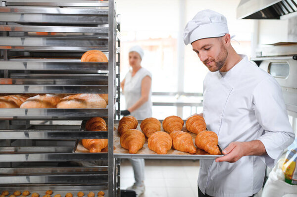 Handsome baker in uniform holding tray full of freshly baked croissants at the manufacturing.