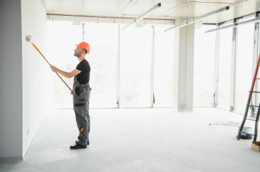 Erkek işçiler apartmanda iç yenileme yapıyorlar. Mutlu gülümseyen, kendine güvenen, beyaz üniformalı, elinde boya silindiri ve tepsisiyle kameraya bakan yetişkin duvar ressamı portresi.