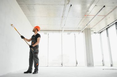 Erkek işçiler apartmanda iç yenileme yapıyorlar. Mutlu gülümseyen, kendine güvenen, beyaz üniformalı, elinde boya silindiri ve tepsisiyle kameraya bakan yetişkin duvar ressamı portresi.