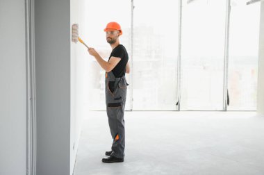 Erkek işçiler apartmanda iç yenileme yapıyorlar. Mutlu gülümseyen, kendine güvenen, beyaz üniformalı, elinde boya silindiri ve tepsisiyle kameraya bakan yetişkin duvar ressamı portresi.