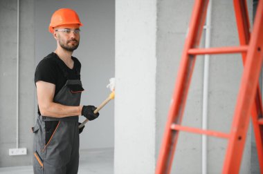 Erkek işçiler apartmanda iç yenileme yapıyorlar. Mutlu gülümseyen, kendine güvenen, beyaz üniformalı, elinde boya silindiri ve tepsisiyle kameraya bakan yetişkin duvar ressamı portresi.