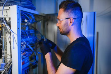 Genç adam sunucu odasında internet ekipmanı ve kablolarıyla çalışıyor..