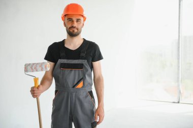 Erkek işçiler apartmanda iç yenileme yapıyorlar. Mutlu gülümseyen, kendine güvenen, beyaz üniformalı, elinde boya silindiri ve tepsisiyle kameraya bakan yetişkin duvar ressamı portresi.