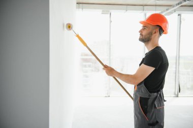 Erkek işçiler apartmanda iç yenileme yapıyorlar. Mutlu gülümseyen, kendine güvenen, beyaz üniformalı, elinde boya silindiri ve tepsisiyle kameraya bakan yetişkin duvar ressamı portresi.