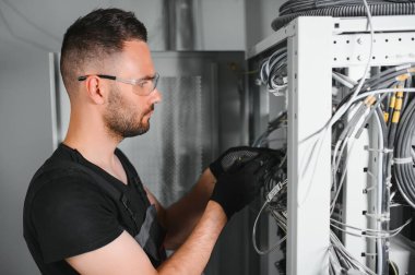 Genç adam sunucu odasında internet ekipmanı ve kablolarıyla çalışıyor..