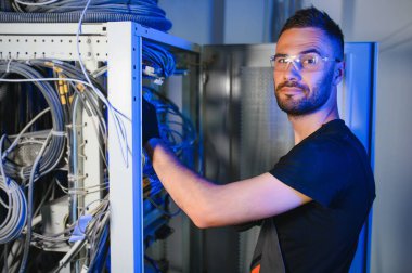 Genç adam sunucu odasında internet ekipmanı ve kablolarıyla çalışıyor..