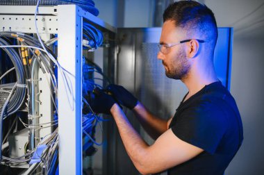 Genç adam sunucu odasında internet ekipmanı ve kablolarıyla çalışıyor..