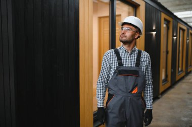 Modüler evlerin inşaat alanında miğfer takmış genç bir marangoz portresi..