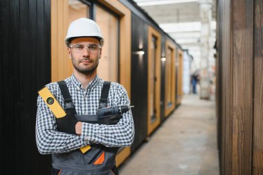 Modüler evlerin inşaat alanında miğfer takmış genç bir marangoz portresi..