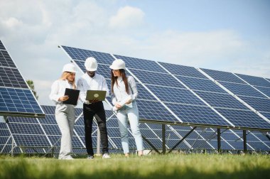 Çok ırklı insanlar ve güneş çiftliğine bakan ve çizimlere bakan güvenlik kaskları. İki mühendis ve teknisyen fotovoltaik inşaat planını açık havada inceliyor..