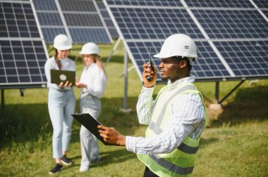 Güneş panellerinin yanında duran mühendislerin portresi. Üç işçi: bir kadın ve iki özel üniformalı adam kameraya bakar ve gülümser..