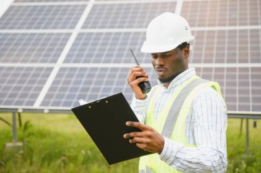 Güvenlik yeleği ve kask giyen Afrikalı inşaat işçisi elinde radyo ve güneş paneli projesiyle arka planda kameraya bakıyor.