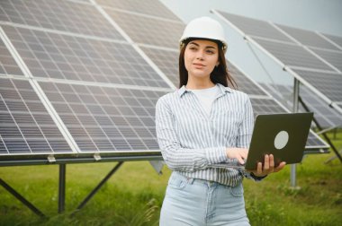 Güneş panellerinin yanında duran genç kadın mühendisin portresi. Beyaz kasklı güzel bir bayan profesyonel kameraya gülümsüyor..
