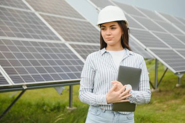 Portrait of electrician engineer in safety helmet and uniform using laptop checking solar panels. Female technician at solar station.