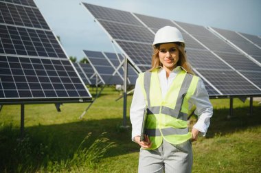 Portrait of electrician engineer in safety helmet and uniform using laptop checking solar panels. Female technician at solar station.