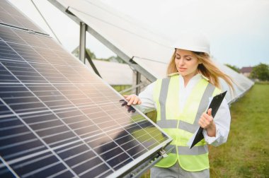 Portrait of electrician engineer in safety helmet and uniform using laptop checking solar panels. Female technician at solar station.