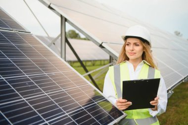Portrait of electrician engineer in safety helmet and uniform using laptop checking solar panels. Female technician at solar station.