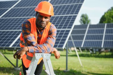 Koruyucu miğfer takmış yakışıklı Afrikalı Amerikalı zanaatkârın portresi. Üniformalı ve aletleri güneş panellerinin arasında duran bir adam..