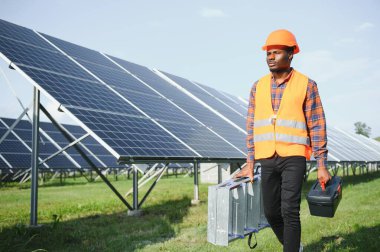 Koruyucu miğfer takmış yakışıklı Afrikalı Amerikalı zanaatkârın portresi. Üniformalı ve aletleri güneş panellerinin arasında duran bir adam..