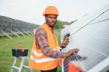 Güneş panellerinde tornavidayla güvenlik kaskı takan Afrikalı Amerikalı bir adam. İstasyonda servis çalışması yaparken araçları kullanan yetkili teknisyen.