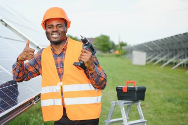 Gri tulumlu ve turuncu başlıklı ehil enerji mühendisi sahada yürürken güneş panellerini kontrol ediyor. Elinde enstrümanlarla pano ve konteynır taşıyan Afrikalı Amerikalı bir adam..