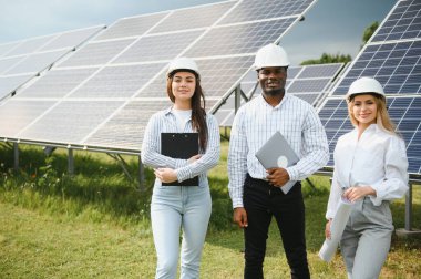 Çok ırklı insanlar ve güneş çiftliğine bakan ve çizimlere bakan güvenlik kaskları. İki mühendis ve teknisyen fotovoltaik inşaat planını açık havada inceliyor..
