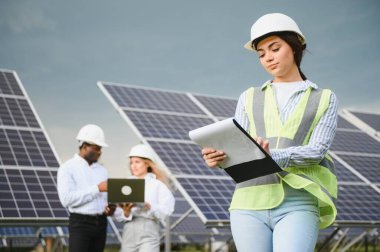 Güneş panellerinin yanında duran genç kadın mühendisin portresi. Beyaz kasklı güzel bir bayan profesyonel kameraya gülümsüyor..