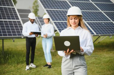 Çok ırklı insanlar ve güneş çiftliğine bakan ve çizimlere bakan güvenlik kaskları. İki mühendis ve teknisyen fotovoltaik inşaat planını açık havada inceliyor..