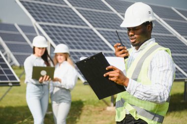 Güneş panellerinin yanında duran mühendislerin portresi. Üç işçi: bir kadın ve iki özel üniformalı adam kameraya bakar ve gülümser..