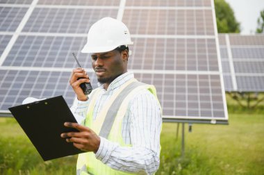 Güvenlik yeleği ve kask giyen Afrikalı inşaat işçisi elinde radyo ve güneş paneli projesiyle arka planda kameraya bakıyor.