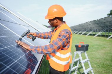 Gri tulumlu ve turuncu başlıklı ehil enerji mühendisi sahada yürürken güneş panellerini kontrol ediyor. Elinde enstrümanlarla pano ve konteynır taşıyan Afrikalı Amerikalı bir adam..