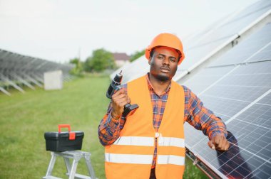 Gri tulumlu ve turuncu başlıklı ehil enerji mühendisi sahada yürürken güneş panellerini kontrol ediyor. Elinde enstrümanlarla pano ve konteynır taşıyan Afrikalı Amerikalı bir adam..