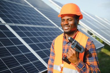 Güneş panellerinde tornavidayla güvenlik kaskı takan Afrikalı Amerikalı bir adam. İstasyonda servis çalışması yaparken araçları kullanan yetkili teknisyen.