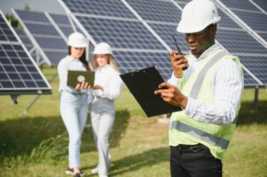 Çok ırklı insanlar ve güneş çiftliğine bakan ve çizimlere bakan güvenlik kaskları. İki mühendis ve teknisyen fotovoltaik inşaat planını açık havada inceliyor..