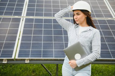 Güneş panellerinin yanında duran genç kadın mühendisin portresi. Beyaz kasklı güzel bir bayan profesyonel kameraya gülümsüyor..