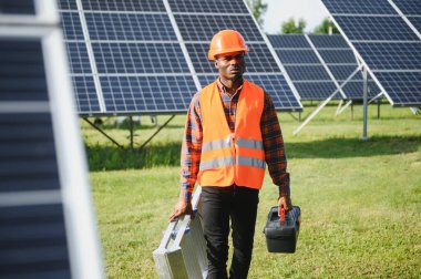 Gri tulumlu ve turuncu başlıklı ehil enerji mühendisi sahada yürürken güneş panellerini kontrol ediyor. Elinde enstrümanlarla pano ve konteynır taşıyan Afrikalı Amerikalı bir adam..