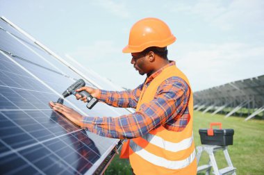 Güneş panellerinde tornavidayla güvenlik kaskı takan Afrikalı Amerikalı bir adam. İstasyonda servis çalışması yaparken araçları kullanan yetkili teknisyen.