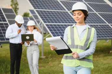 Çok ırklı insanlar ve güneş çiftliğine bakan ve çizimlere bakan güvenlik kaskları. İki mühendis ve teknisyen fotovoltaik inşaat planını açık havada inceliyor..