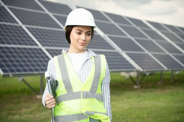 Güneş panellerinin yanında duran genç kadın mühendisin portresi. Beyaz kasklı güzel bir bayan profesyonel kameraya gülümsüyor..