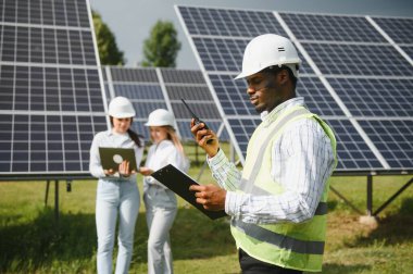Çok ırklı insanlar ve güneş çiftliğine bakan ve çizimlere bakan güvenlik kaskları. İki mühendis ve teknisyen fotovoltaik inşaat planını açık havada inceliyor..